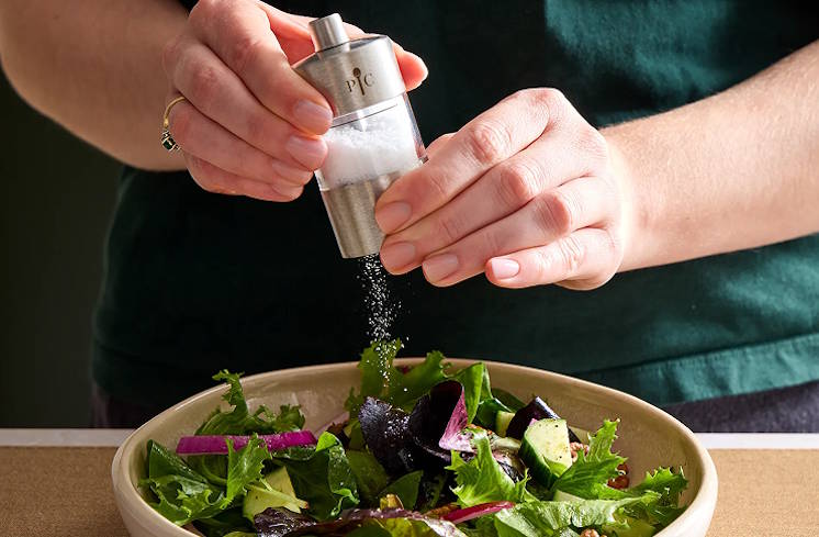 A woman sitting on a chair, shopping Pampered Chef on her phone; with a pop-up display from the phone demonstrated in the background reading: what customers are loving. ready to start shopping? See what's popular right now or shop all products.
